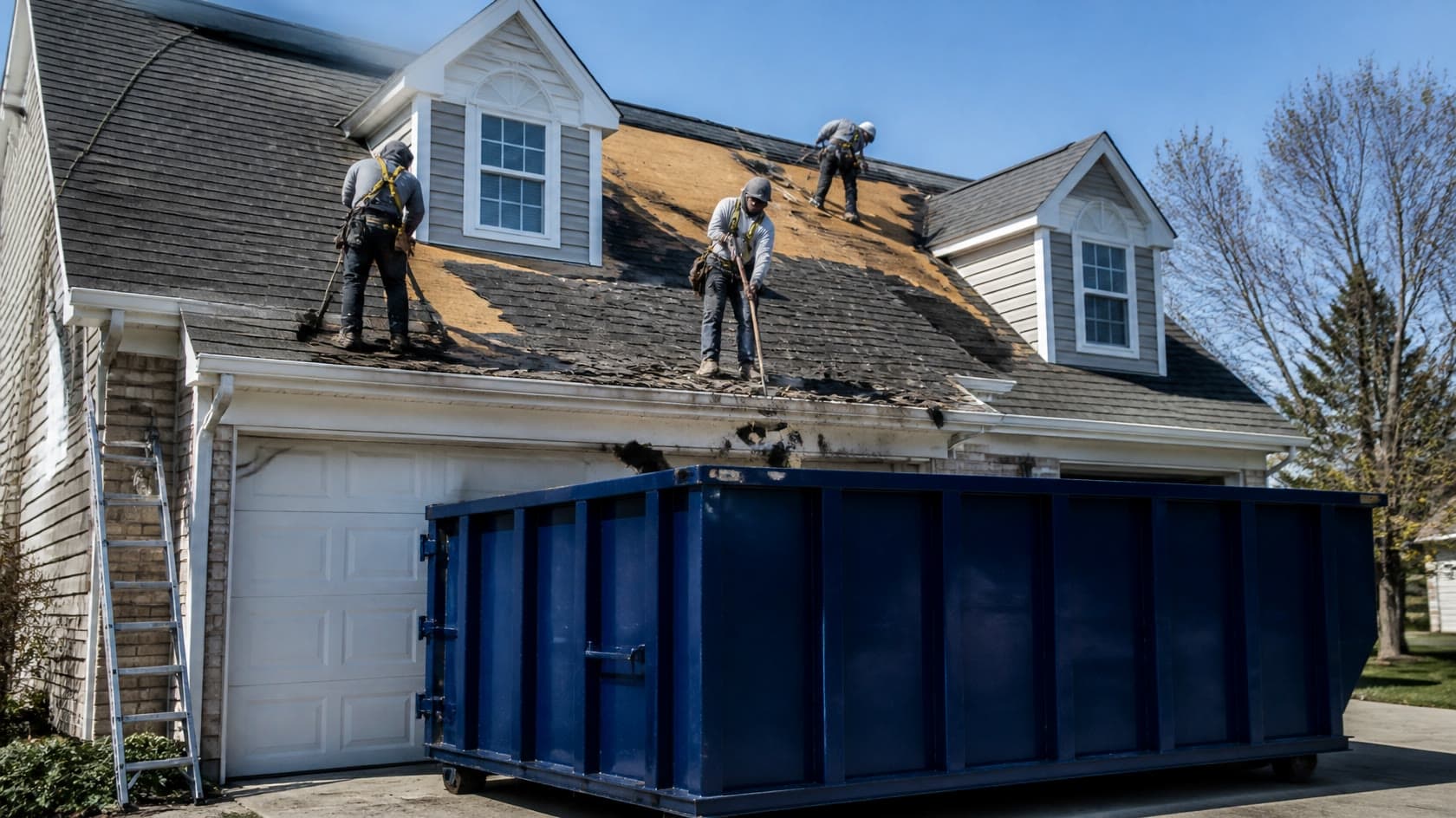 Roofing crew tearing off old shingles from a Michigan home with a navy blue roll-off dumpster in the driveway